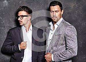 Handsome fashion male models men dressed in elegant suits posing near dark gray wall