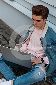 Handsome fashion young freelancer man in stylish denim casual jeans clothes sits with laptop and works remotely on creative