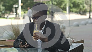 Handsome fashionable afro american business man stirring juice while usinh smartphone in outside cafe