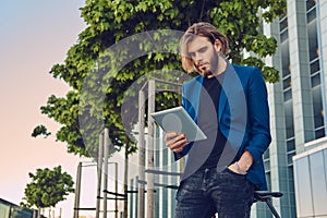 A handsome fashionable bearded male with long hair dressed in stylish clothes, working with a tablet computer after