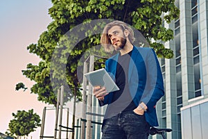 A handsome fashionable bearded male with long hair dressed in stylish clothes, working with a tablet computer after