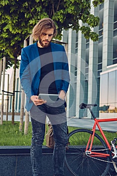 A handsome fashionable bearded male with long hair dressed in stylish clothes, working with a tablet computer after