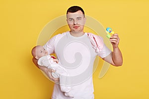 Handsome father and baby child posing isolated over yellow background, dad holds beanbag in hands, wearing casual white t shirt,