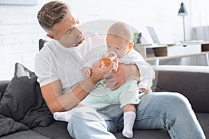 handsome father sitting on couch and feeding baby daughter from bottle