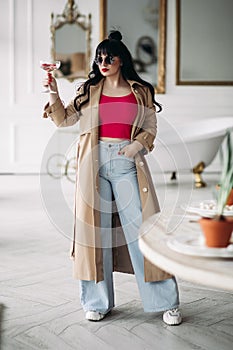 young caucasian woman with long dark wavy hair in fashionable clothes poses for the camera with a glass of wine or juice