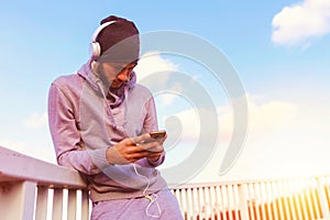 Handsome fit man taking a break after intensive training outdoors, texting on his smart phone wearing headphones
