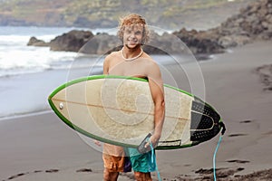 Handsome fit young blond man with mock up surfboard waits for wave to surf spot at sea ocean beach with black sand and