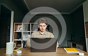 Handsome freelance programmer in a shirt and wireless headphones works on a laptop at home in a cozy bedroom, looks intently at