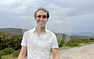 a handsome French or Spaniard man in a white shirt stands against the backdrop of mountains photo