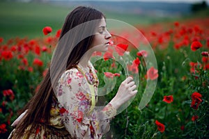 A handsome girl with long hair and natural skin, standing in a fiel of red poppies and holding a red poppy in hands