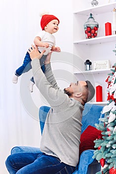 Handsome dad throwing up his amusing infant boy in the air near festive decorated christmas tree.