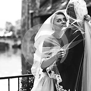 Handsome groom and beautiful bride kissing under veil in old street closeup b&w