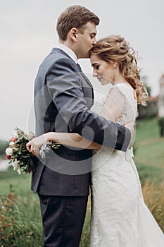 Handsome groom embracing beautiful bride and kissing her on the forehead, happy newlyweds portrait, couple hugging outdoors