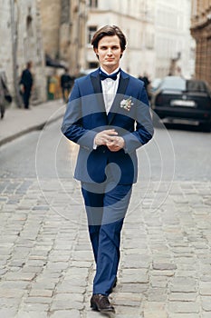 Handsome groom portrait, smiling groom in elegant blue suit with boutonniere posing outdoors close-up, man wedding fashion concept