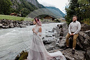 handsome groom sitting on rock and looking at beautiful tender bride standing near mountain river