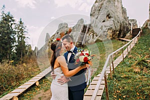 Handsome groom in stylish blue suit kissing his white dressed bride holding bouquet of roses on majestic mountain