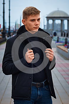 Handsome guy in a black jacket with a fur collar against the background of the city embankment with a rotunda in blur.