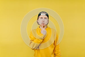 A handsome guy in a cap and a yellow sweatshirt stands on a yellow background and looks up with a pensive face