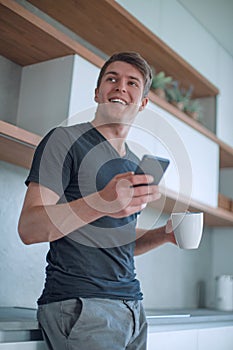 Handsome guy with a Cup of coffee and a smartphone standing in the kitchen