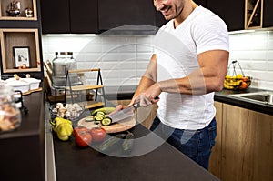 Handsome guy cutting a cucumber with a knife in the kitchen as he prepares a vegetable dish