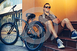 Handsome Guy Cyclist In Sunglasses With Backpack Sitting On The Steps Near The Bicycle Bike And Resting