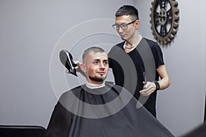 Handsome guy has a haircut in a barbershop, a Kazakh barber dries a client`s hair dryer against a gray wall, a hairdresser makes