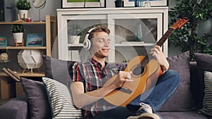 Handsome guy melomaniac is listening to music in headphones and playing the guitar relaxing at home on sofa in leisure