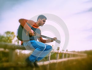 Handsome guy playing guitar sitting on wooden fence in nature.