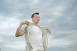 Handsome guy puts on a shirt against the background of the sky. Men`s fashion, unity with nature