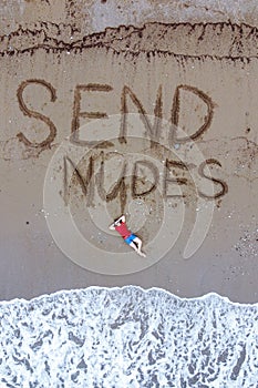 Handsome guy in red t-shirt and blue male boxers is lying on beach washed by foaming sea waves. send nudes inscription on wet sand