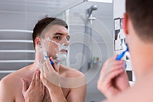 Handsome guy shaving his face with a razer and looking at the mi