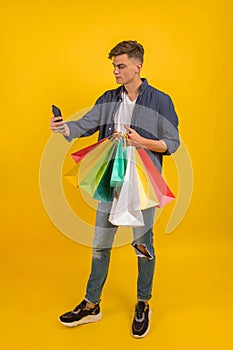 Handsome guy with shopping bags is using a mobile phone and smiling. Portrait of a smiling man holding shopping bag over yellow ba