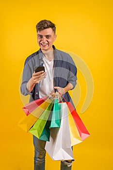 Handsome guy with shopping bags is using a mobile phone and smiling. Portrait of a smiling man holding shopping bag over yellow ba