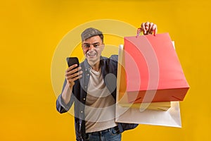 Handsome guy with shopping bags is using a mobile phone and smiling. Portrait of a smiling man holding shopping bag over yellow ba