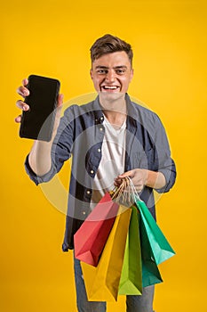 Handsome guy with shopping bags is using a mobile phone and smiling. Portrait of a smiling man holding shopping bag over yellow ba