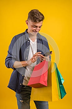 Handsome guy with shopping bags is using a mobile phone and smiling. Portrait of a smiling man holding shopping bag over yellow ba