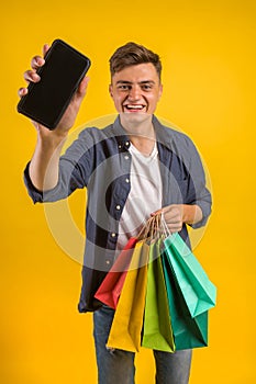 Handsome guy with shopping bags is using a mobile phone and smiling. Portrait of a smiling man holding shopping bag over yellow ba