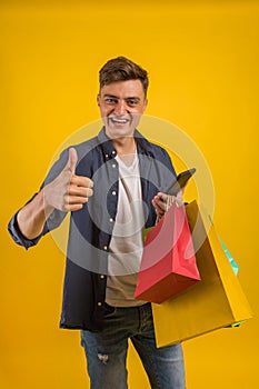 Handsome guy with shopping bags is using a mobile phone and smiling. Portrait of a smiling man holding shopping bag over yellow ba