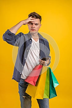 Handsome guy with shopping bags is using a mobile phone and smiling. Portrait of a smiling man holding shopping bag over yellow ba