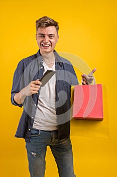 Handsome guy with shopping bags is using a mobile phone and smiling. Portrait of a smiling man holding shopping bag over yellow ba