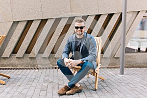 Handsome guy is sitting on chair outdoor on cafe background. He wears gray jacket, jeans, brown shoes, sunglasses. He is