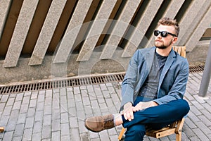 Handsome guy is sitting on chair outdoor on concrete wall background. He wears gray jacket, jeans, sunglasses