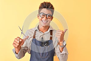 Handsome hairdresser man wearing apron holding scissors over isolated yellow background pointing and showing with thumb up to the