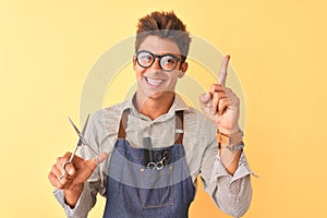 Handsome hairdresser man wearing apron holding scissors over isolated yellow background very happy pointing with hand and finger