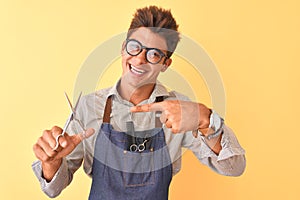 Handsome hairdresser man wearing apron holding scissors over isolated yellow background very happy pointing with hand and finger