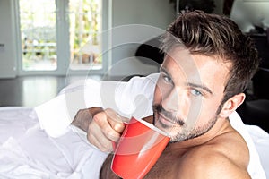 Handsome hairy naked muscular man with beard sixpack abs lying in bed covered with sheet drinking coffee