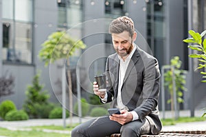 Handsome happy bearded businessman sitting on bench with cup coffee use mobile phone browse smartphone in hands reads good news