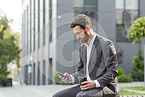 Handsome happy bearded businessman sitting on bench with cup coffee use mobile phone browse smartphone in hands reads good news