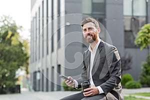 Handsome happy bearded businessman sitting on bench with cup coffee use mobile phone browse smartphone in hands reads good news