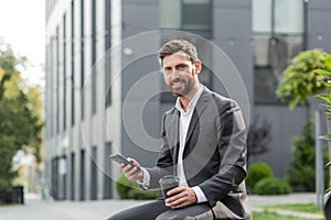 Handsome happy bearded businessman sitting on bench with cup coffee use mobile phone browse smartphone in hands reads good news